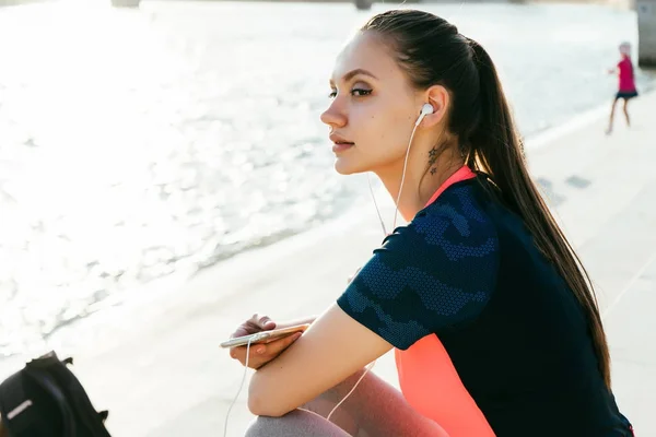 Jovem fitness menina em sportswear descansando depois de correr pelo rio, ouvindo música em fones de ouvido — Fotografia de Stock