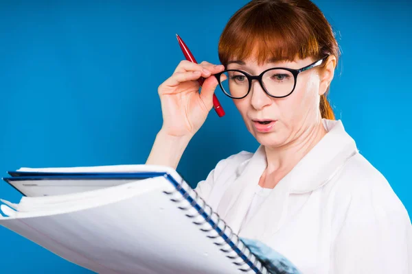 Die erwachsene rothaarige Ärztin mit Brille und weißem Arztkittel blickt überrascht in ihr Notizbuch. — Stockfoto