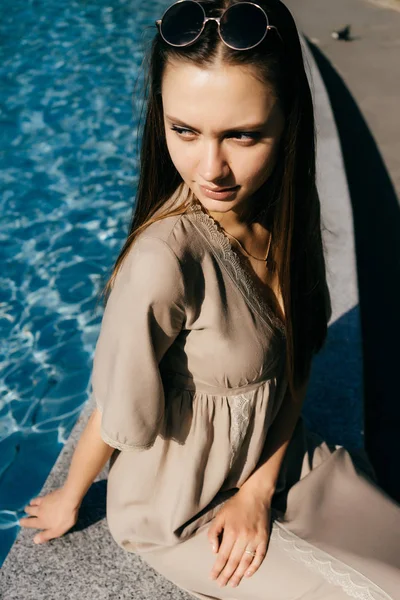 Menina encantadora em um vestido bege elegante senta-se à beira da piscina com água azul, goza do sol — Fotografia de Stock