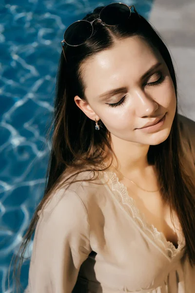 Chica joven atractiva disfruta del sol, se sienta junto a la piscina con agua azul, sus ojos están cerrados — Foto de Stock