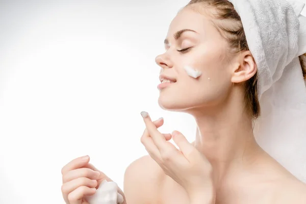 A beautiful young girl looks after her face, puts a cream on her face, on her head a white towel — Stock Photo, Image