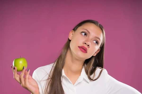 Trött ung flicka i vit skjorta håller ett grönt äpple, på en rosa bakgrund — Stockfoto