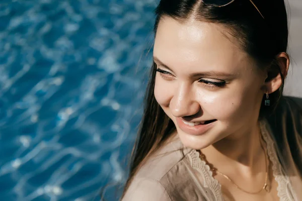 Menina bonita atraente em um vestido bege posando à beira da piscina com água azul, apreciando o sol e rindo — Fotografia de Stock