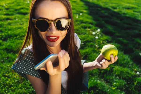 Žena mluvící hlasové zprávy na smartphone, sedí na zelené trávě — Stock fotografie