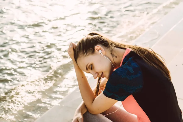 Linda joven en ropa deportiva descansando después de trotar junto al río al atardecer, escuchando música en los auriculares — Foto de Stock