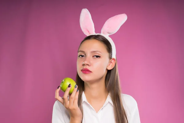 Una giovane ragazza triste con una camicia bianca, con un orlo a forma di orecchie di coniglio in testa, tiene in mano una mela verde — Foto Stock