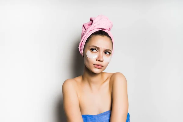 Νεαρό κορίτσι με μια ροζ πετσέτα στο κεφάλι της, ψάχνει προσεκτικά επάνω, κάτω από τα μάτια των patches με σιλικόνη — Φωτογραφία Αρχείου