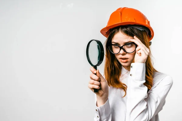 En flicka med glasögon innehar en orange byggarbetarhjälm med sin hand och ser ut genom ett förstoringsglas — Stockfoto