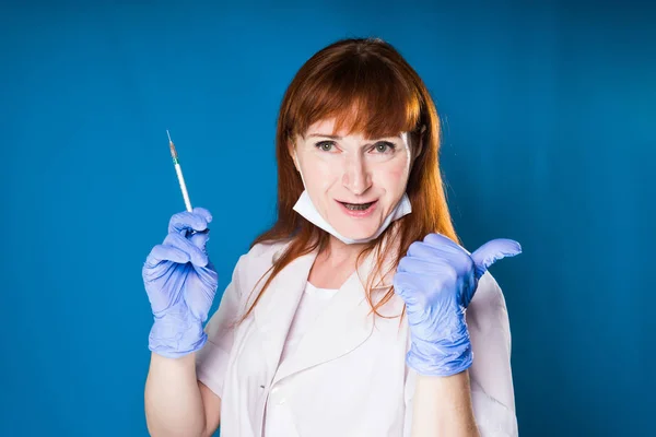 happy red-headed nurse wearing a mask and gloves going to make an injection and showing a thumbs up, isolated on a blue background