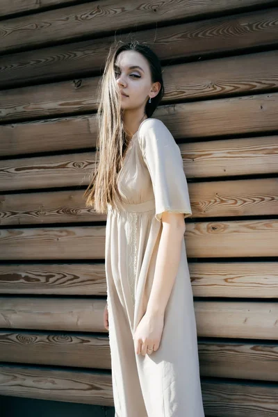 Menina bonito em um vestido bege longo está em pé junto à parede de madeira e olha para a câmera — Fotografia de Stock