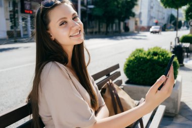 güzel bej elbise uzun saçlı kız gülümseyerek ahşap bir bankta oturur bir akıllı telefon tutar