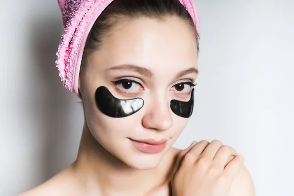 Красивая молодая девушка ухаживает за кожей лица, под глазами черные силиконовые пятна, на голове розовое полотенце — стоковое фото