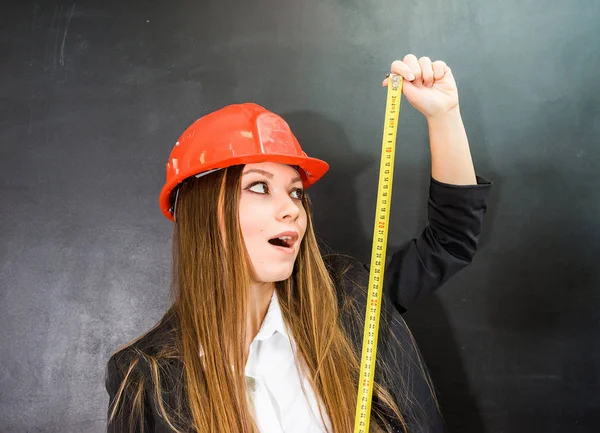 Jovem construtor menina surpreso em um capacete laranja protetora mantém uma fita métrica na mão — Fotografia de Stock