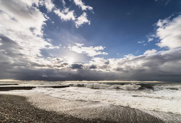Raging sea near the beach from the pebbles, the sky is covered with gray clouds, the sun is almost invisible — Stock Photo, Image