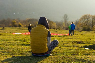 bir ceket ve başlık bir açıklıkta yeşil çimenlerin üzerinde oturan insanlar ve dağlar bakarak bir adam