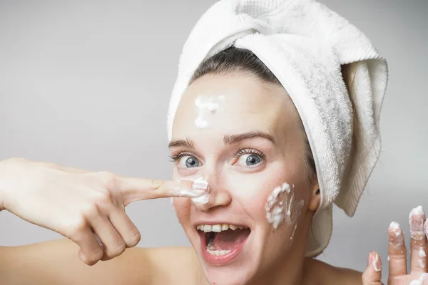 Mujer divertida en una toalla en la cabeza feliz limpia la piel con espuma sobre un fondo blanco aislado. Concepto de limpieza del cuidado de la piel —  Fotos de Stock