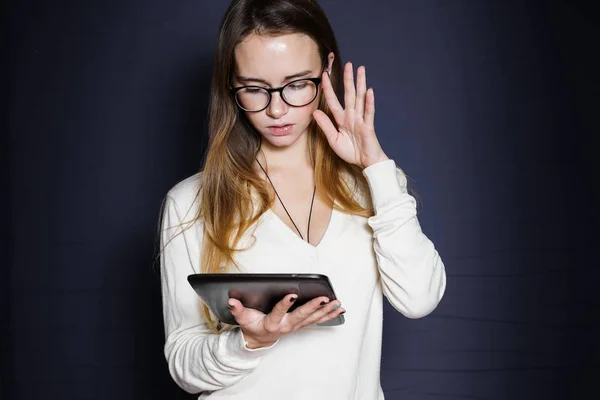 Langharige jong meisje in een witte jas en bril kijkt naar de tablet — Stockfoto