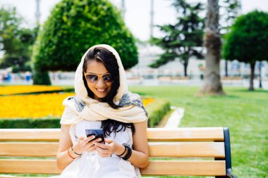 mutlu esmer kadın güneş gözlüğü ve parkta bankta oturmuş, gülümseyen ve onun smartphone bakarak başörtüsü ile