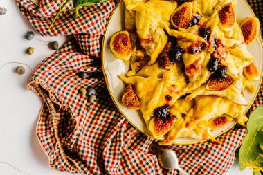 Beyaz bir masada işte damalı bir masa örtüsü, bir tabak sıcak hamur işleri ve tatlı incir, yeşil yaprakları