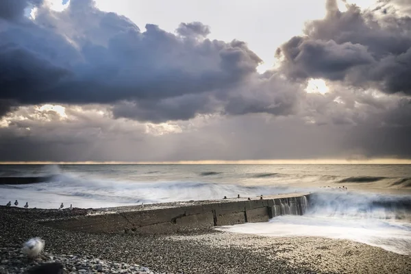 The sky is covered with gray clouds, the sun is almost invisible, the white waves near the shore — Stock Photo, Image