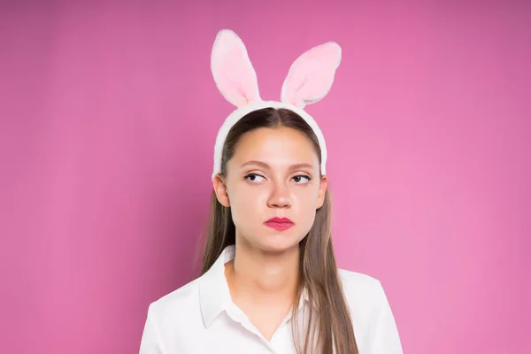 Verdrietig meisje in een wit overhemd, in een rand in de vorm van Haas oren op haar hoofd poseren op een roze achtergrond — Stockfoto