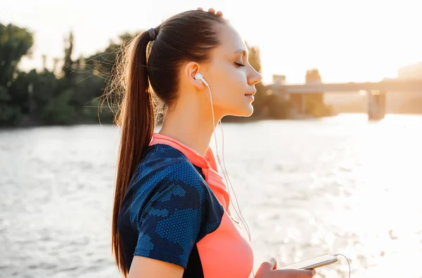 Schöne Fitness-Frau in Sportbekleidung, die bei Sonnenuntergang Musik über Kopfhörer am Fluss hört — Stockfoto
