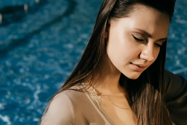 Очаровательная красивая девушка с длинными волосами сидя у бассейна с голубой водой, позируя на солнце, ее глаза закрыты — стоковое фото