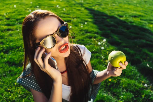 Mujer enojada hablando con móvil, sostiene una manzana en su mano, se sienta en la hierba verde en el parque — Foto de Stock