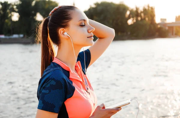 Glad ung fitness flicka i sportkläder som lyssnar på musik i hörlurar, njuta av solen vid floden — Stockfoto