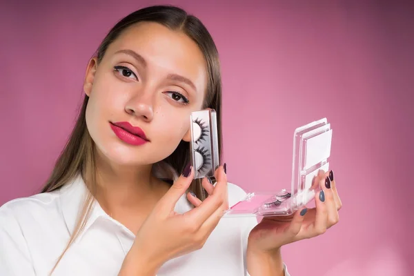 Belle fille aux cheveux longs dans une chemise blanche tient un paquet enveloppé avec de faux cils, sur un fond rose — Photo