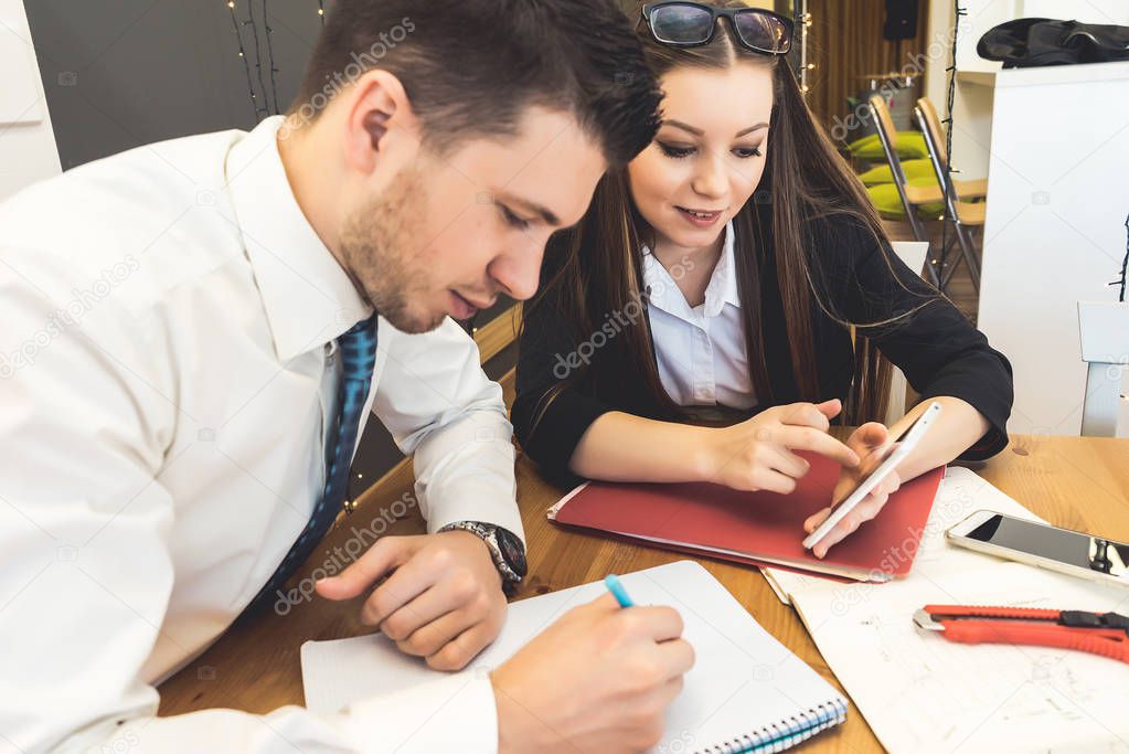 The lady and the man are sitting at the desk in the office and working. A young man writes something in a notebook, a girl looks at the phone