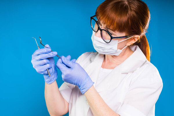 adult red-haired woman doctor in glasses with a protective mask and white coat holds in hands a medical dental instruments