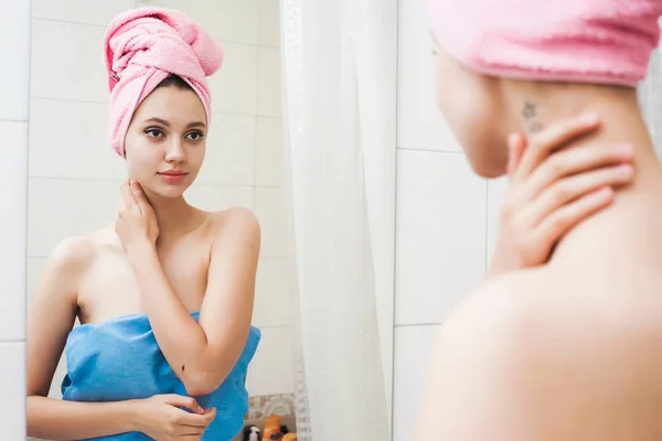 Красивая молодая девушка с чистой кожей, с розовым полотенцем на голове, глядя на себя в зеркало — стоковое фото