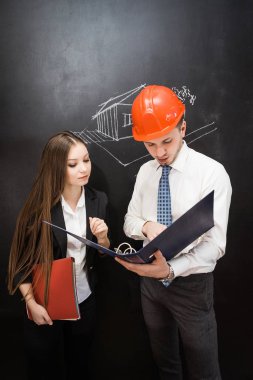 genç bir adam bir portakal inşaat kask içinde bir klasör elinde tutar ve kız bir plan-proje gösterir. Bir ev düzeni tahtada çizilir