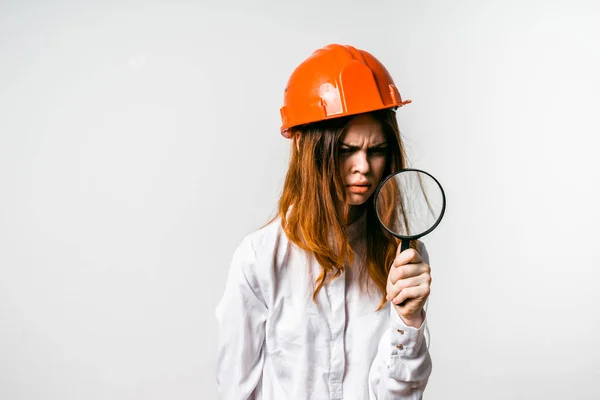 Uma menina em uma camisa branca e em um capacete de construção laranja olha através da lupa com um olhar irritado — Fotografia de Stock