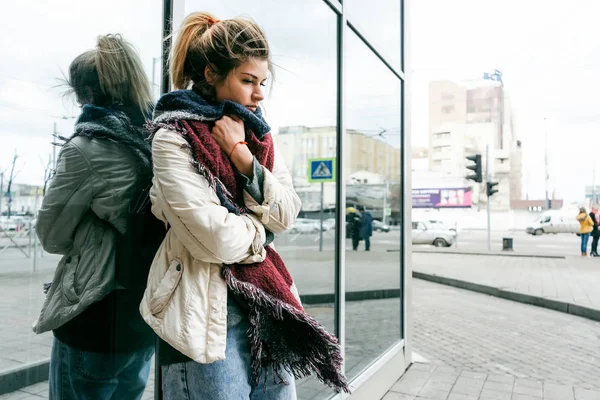 A girl wrapped in a warm scarf stands on a city street leaning against the wall of the building and looks down. the girl froze — Stock Photo, Image