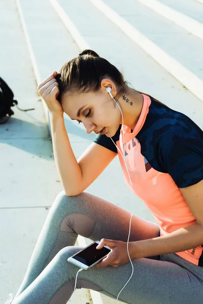 Flicka i sport kostym sitter på trappan och lyssnar på musik i hörlurar — Stockfoto