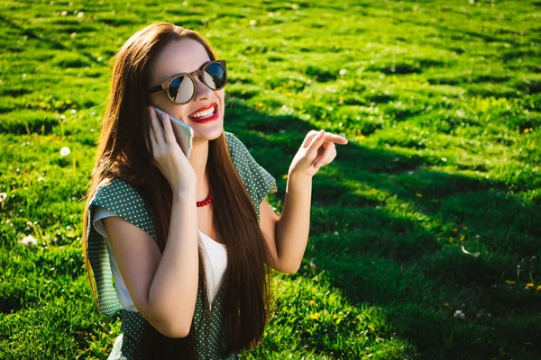 Happy woman in glasses pointing to something on grass,uses mobile — Stock Photo, Image
