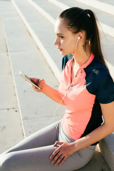 the girl sat down on the steps on the riverbank to listen to the music in the headphones after training. a woman is holding a phone in her hands and looking into the distance