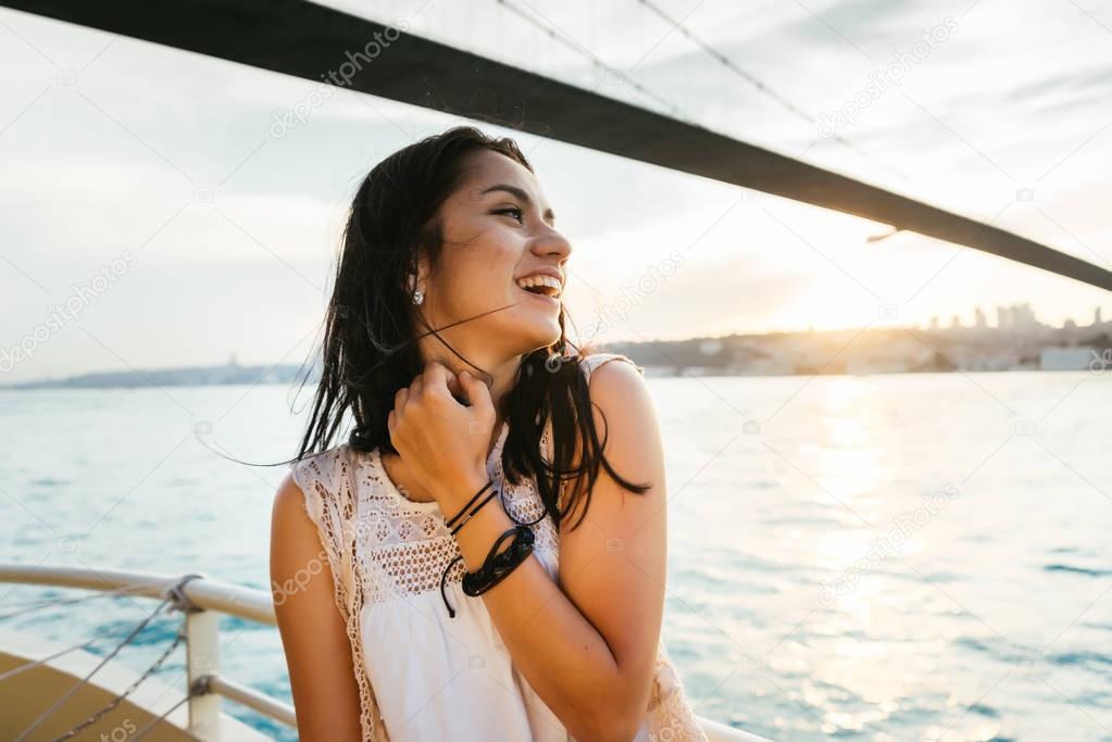 A beautiful Latin girl on a yacht sails under a modern bridge at sunset and laughs