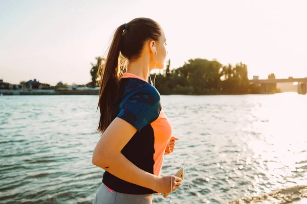 Uma menina em um terno esportivo corre ao longo da orla e ouve música com fones de ouvido — Fotografia de Stock