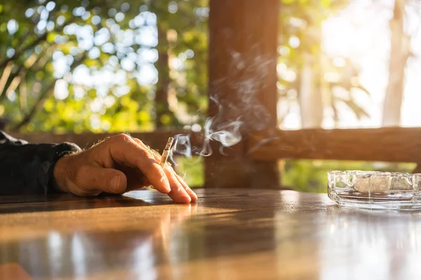 Egy ember kéz tartja a cigarettát. Füstölög a cigaretta, a hamutartót van az asztalon. kívül az ablak meleg napos idő — Stock Fotó