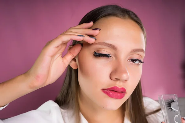 Flicka med stora läppar limmade på ena ögat lösögonfransar, isolerad på en rosa bakgrund — Stockfoto