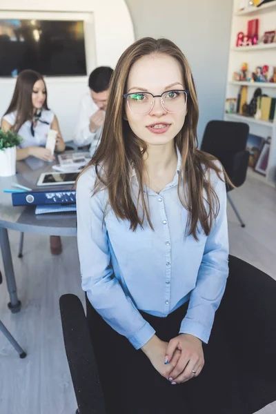 Schönes junges erfolgreiches Mädchen, Büroangestellte, Brille tragend, in die Kamera schauend, lächelnd, hinter ihr ein Mann und eine Frau, die zusammen arbeiten — Stockfoto
