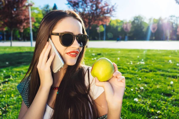 Pozitivní žena s úsměvem a těší slunce, v brýlích, mluvil po telefonu a v ruce držel zelené jablko — Stock fotografie