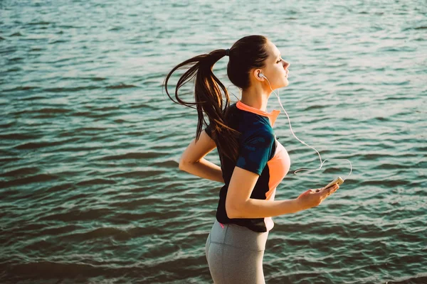 Giovane fitness girl che fa jogging al fiume, ascoltando musica con le cuffie — Foto Stock