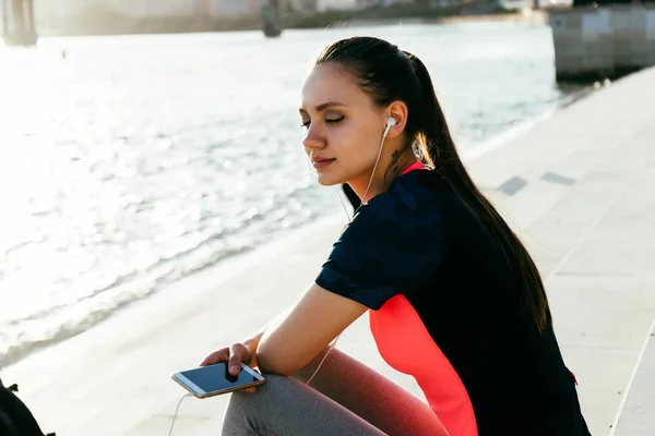 ヘッドフォンで音楽を聴き、川でトレーニングの後休んでいる若い美しいフィットネス女の子 — ストック写真