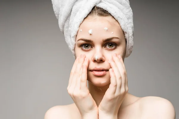 Encantadora jovem com uma toalha branca na cabeça cuida de seu rosto, aplica creme , — Fotografia de Stock
