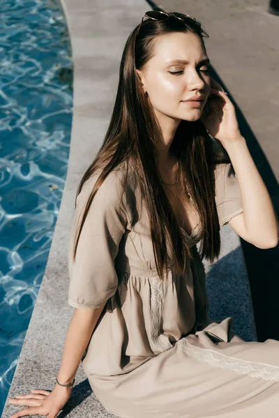 Ελκυστικές όμορφο κορίτσι μοντέλο κάθεται δίπλα στην πισίνα με το μπλε νερό, απολαμβάνοντας τον ήλιο, τα μάτια κλειστά — Φωτογραφία Αρχείου