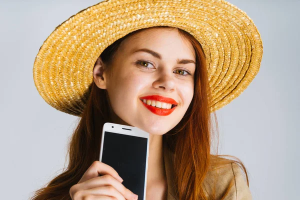 Gelukkig aantrekkelijk meisje met rode lippenstift, in een stro hoed houdt een smartphone in haar hand, glimlachend — Stockfoto